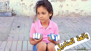 طفلة تبتسم وهي تبيع المناديل على الرصيف، ترتدي ملابس بسيطة وعينيها تحملان براءة وقوة.